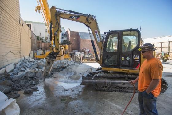 Worker sprays water on construction site while excavator removes concrete debris at Powell Street.