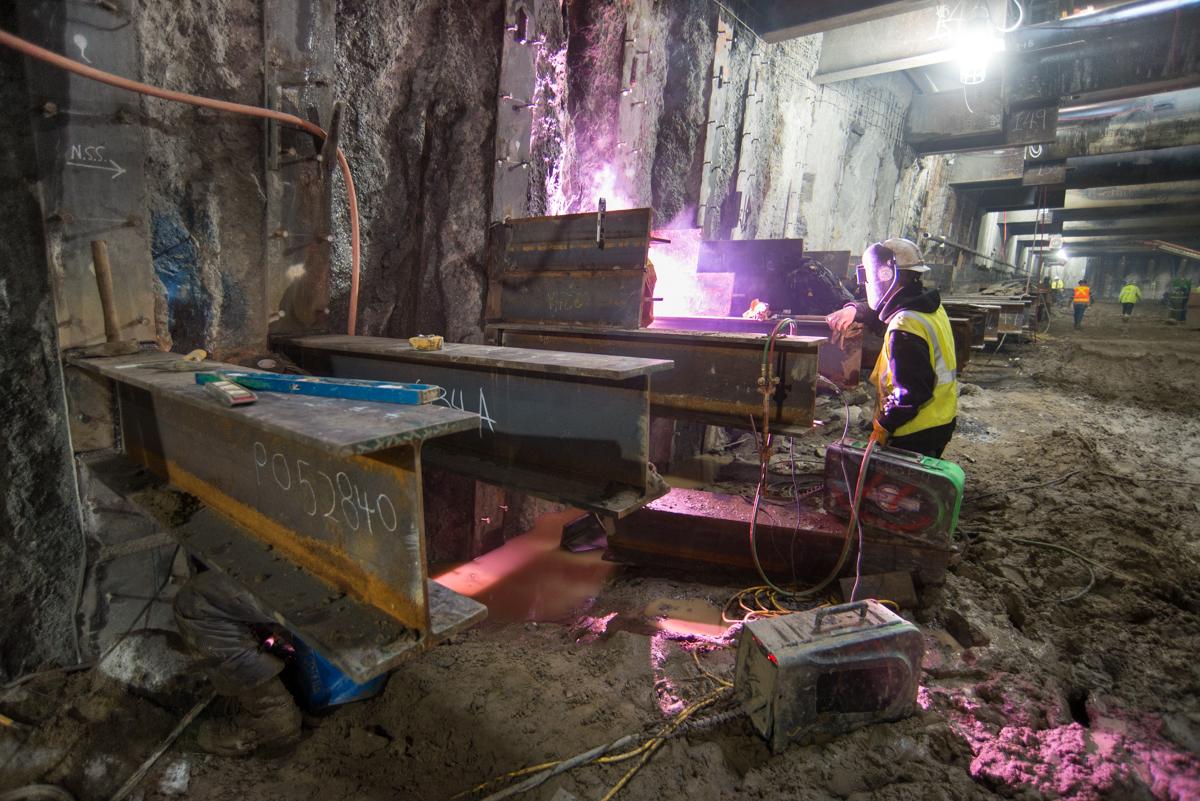 Worker in safety vest welding steel brackets for temporary bracing during underground transit station excavation.