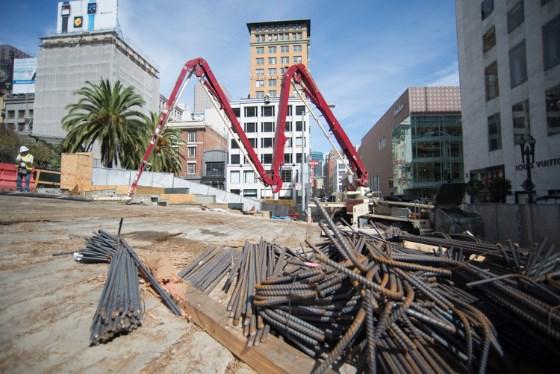 Construction site with red concrete pump boom, steel reinforcement bars, and downtown San Francisco buildings.