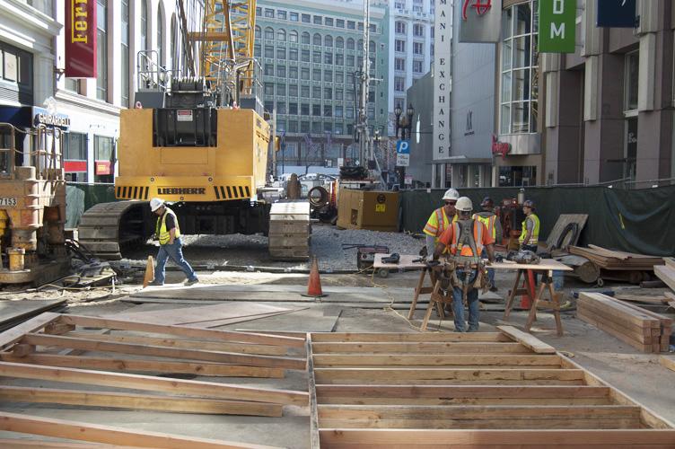 Construction crew installing subsurface walls at Union Square/Market Street Station site with heavy equipment and wooden framing.
