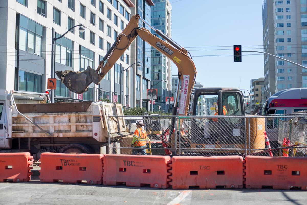 Excavator loads soil into dump truck at urban construction site surrounded by red barriers and high-rise buildings.