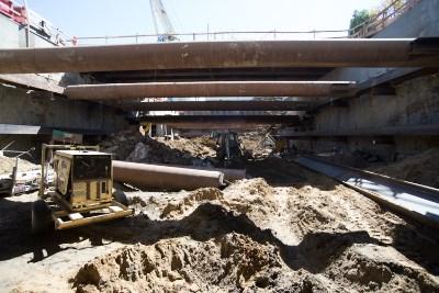 Construction excavation beneath concrete bridge structure with steel support beams and utility pipes visible.