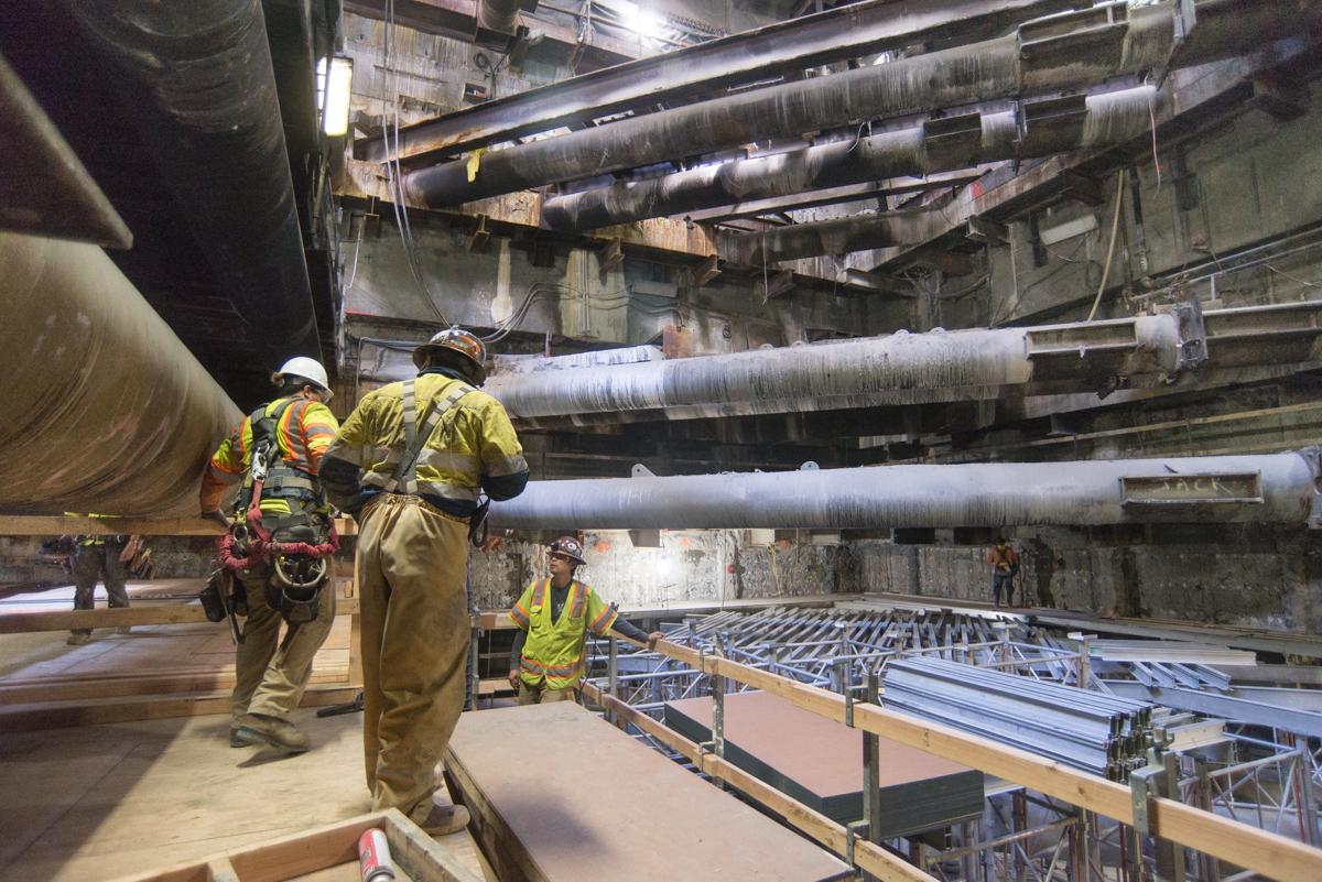Workers in safety gear construct rebar cages inside a transit station headhouse under large overhead pipes.