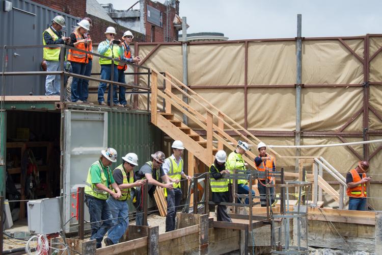Construction workers in hard hats and safety vests on wooden scaffolding and platforms at an active construction site with industrial buildings in background.