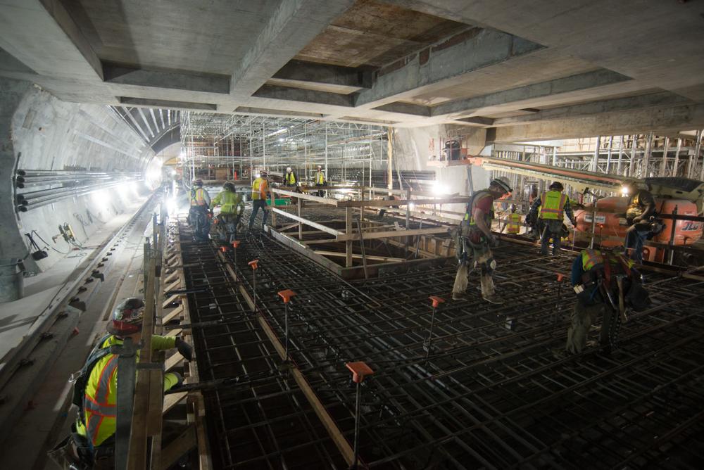 Workers in safety vests install rebar reinforcement on an underground transit station platform under concrete ceiling beams.