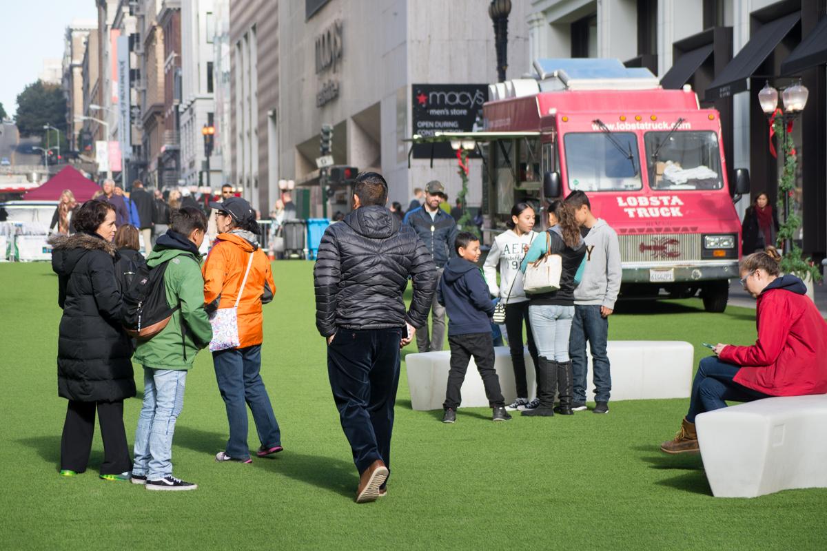 People gather on artificial green turf in an urban plaza with a red food truck and city buildings in the background.