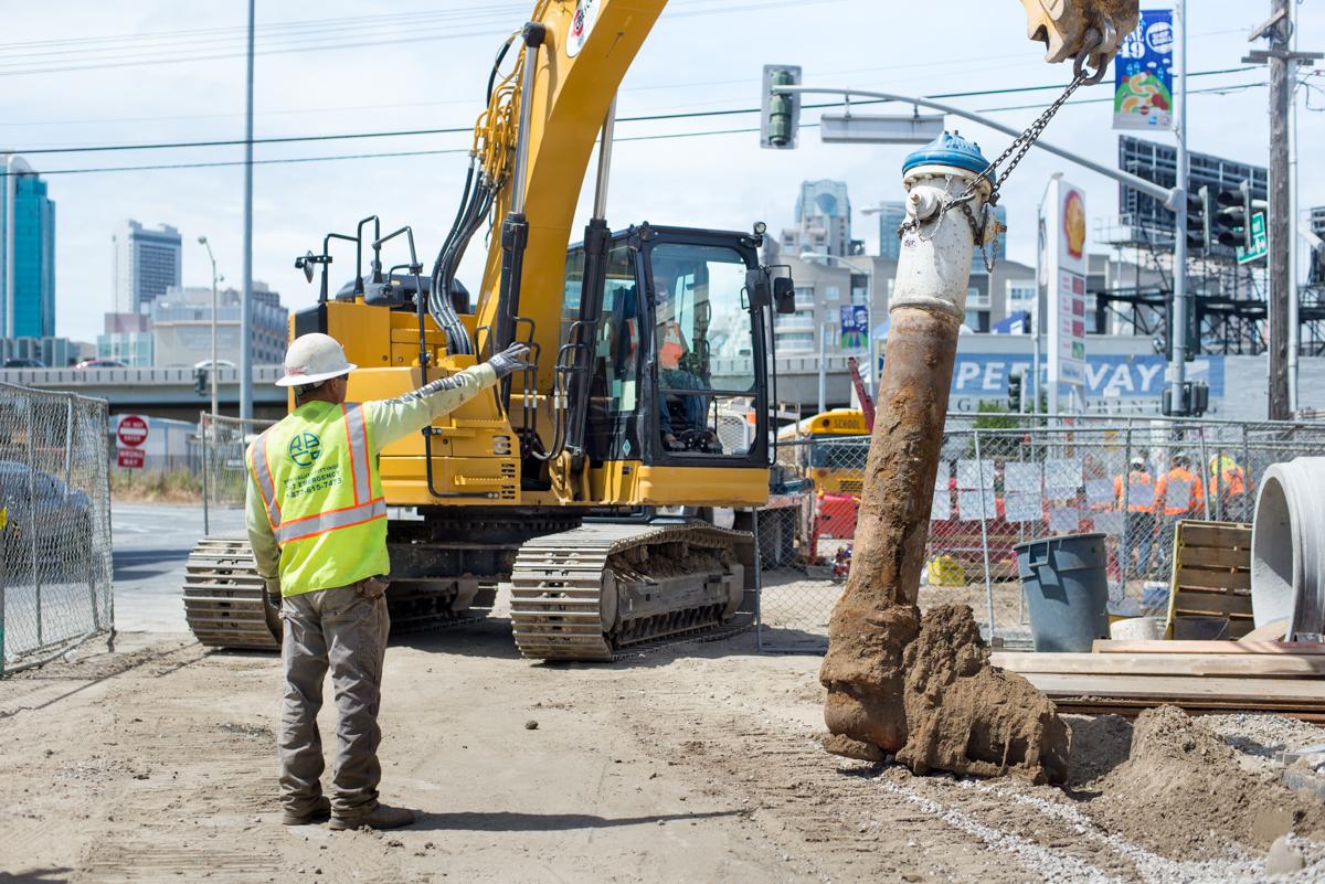 Worker in safety vest directs excavator operator lowering old utility hydrant at construction site.