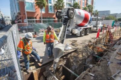 Construction workers direct concrete mixer truck to pour into underground utility trench at urban work site.