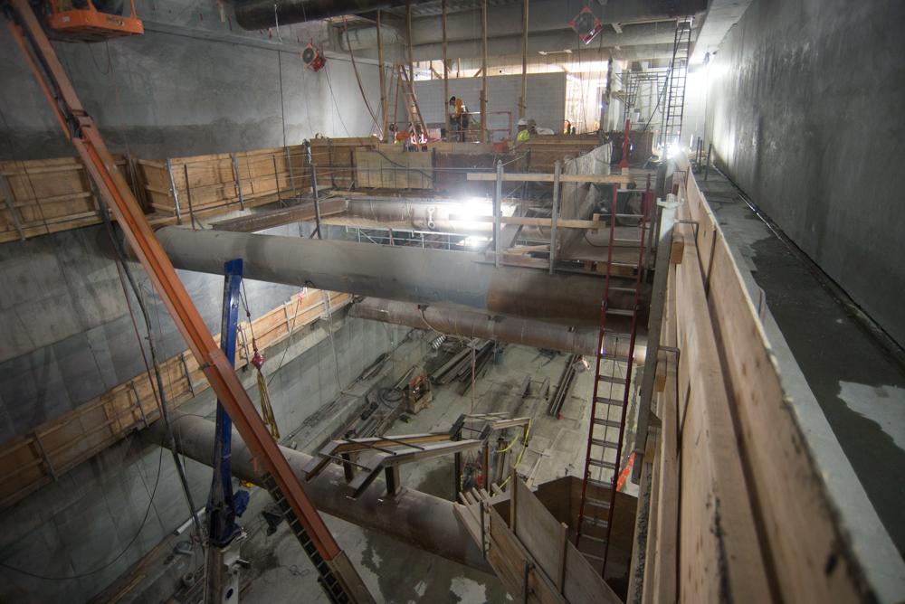 Construction site showing underground transit station with workers on mezzanine level and platform below installing structural elements and steel framing.