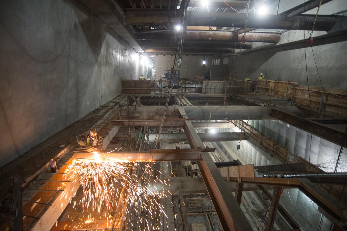 Worker welding steel beam during underground transit construction with concrete walls and overhead lighting.