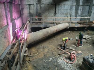 Workers welding brackets on large steel I-beam in underground construction site with concrete walls.