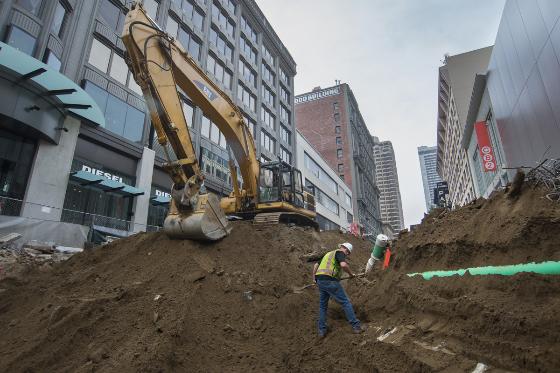 Construction worker in safety vest stands on dirt pile next to yellow excavator on urban street with buildings.