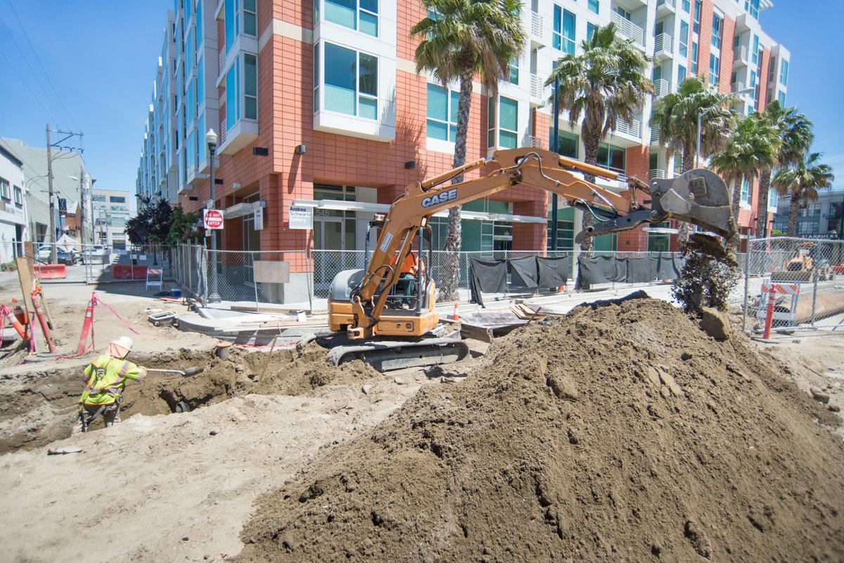 Construction excavator and worker performing trench excavation on urban street with residential building in background.