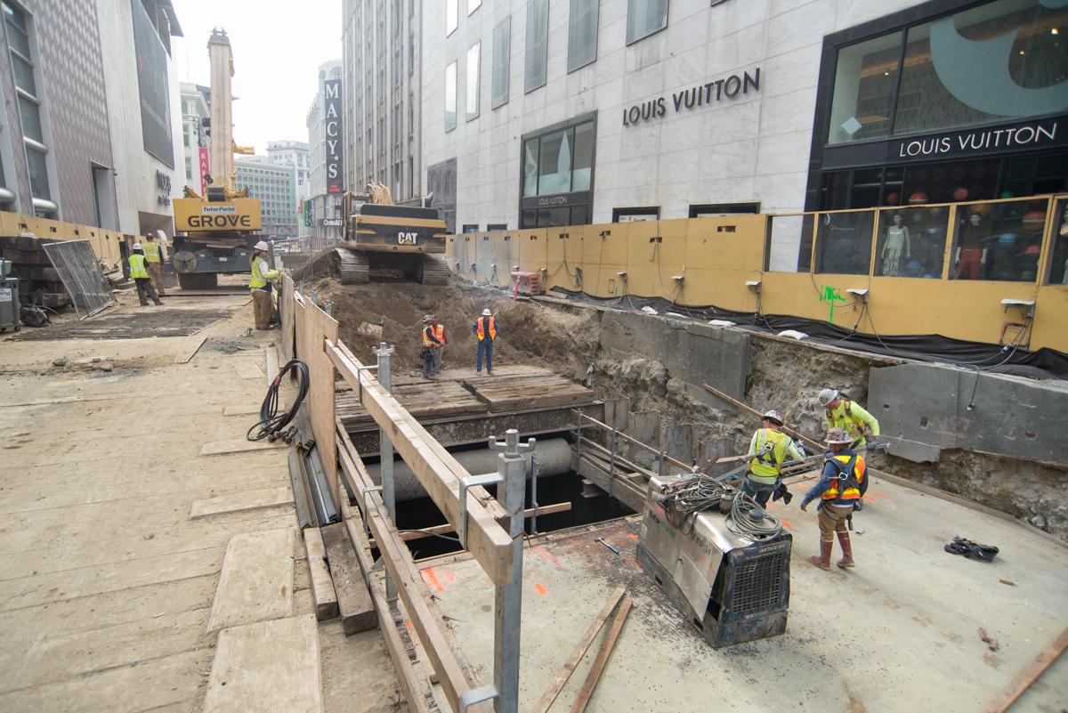 Construction crew working in underground access shaft on downtown street with heavy equipment and safety barriers.
