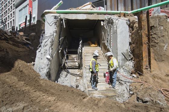 Two workers in safety vests stand at the entrance of an underground transit tunnel under construction at Ellis and Stockton streets.