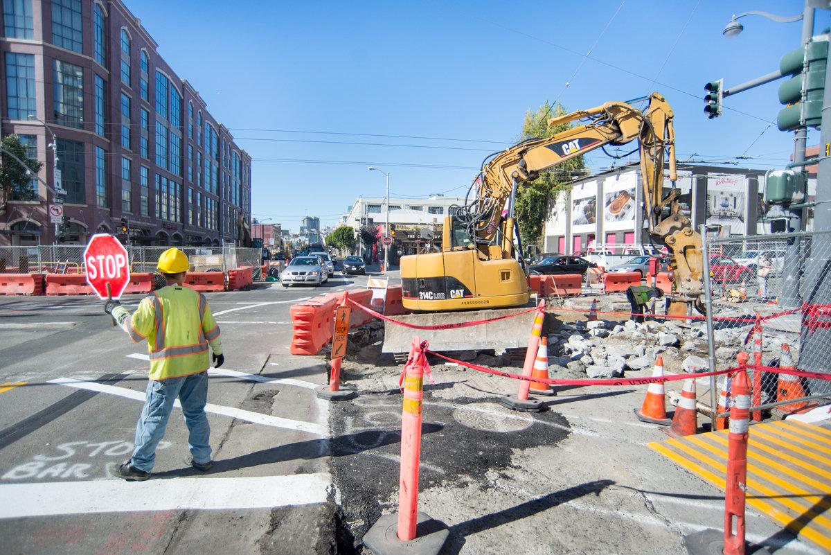 Construction worker with stop sign directs pedestrians as excavator breaks up pavement on city street.