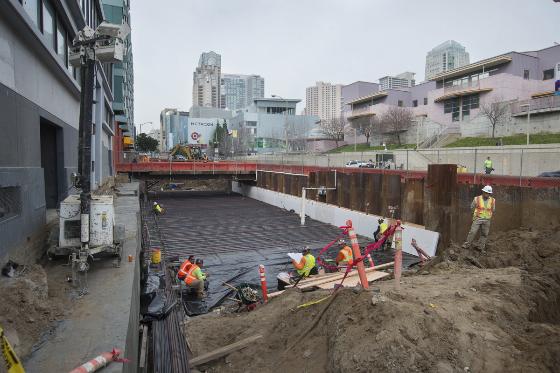 Workers in safety gear install waterproofing on 4th Street construction site with city skyline in background.