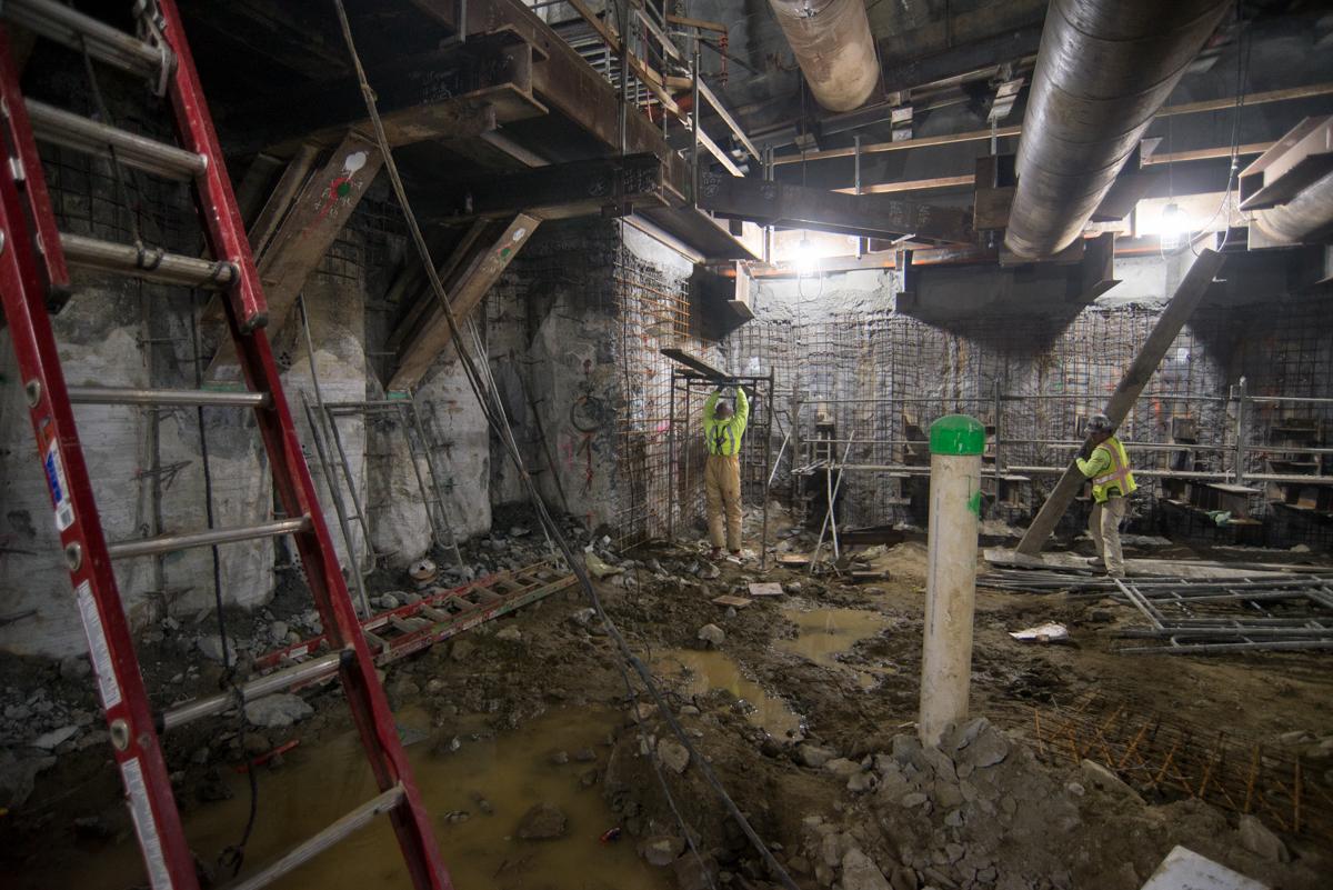Construction workers in safety vests work in muddy underground station box with exposed concrete walls and metal scaffolding.