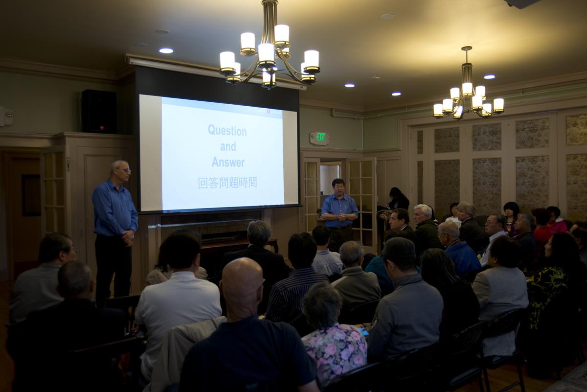 Community meeting with audience seated facing projection screen displaying "Question and Answer" text, with two presenters in blue shirts standing at front of room.