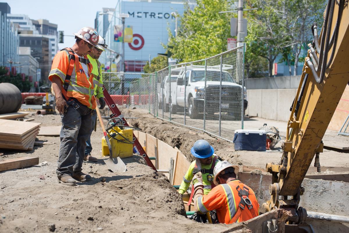 Construction workers excavate and install utilities at transit station site with heavy equipment and safety barriers.