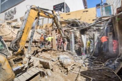 Yellow excavator demolishing Union Square Garage southeast corner with workers and debris visible on urban street.
