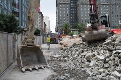 Excavator with bucket sorting demolished concrete and rubble on urban street surrounded by tall buildings.
