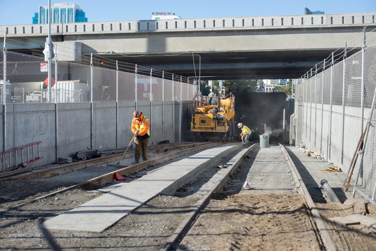 Construction workers install track crossovers in light rail tunnel portal with yellow machinery and concrete work visible.