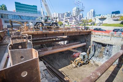 Construction site with excavation pit, slurry wall, jackhammer operator, and dust control water spray in urban setting.