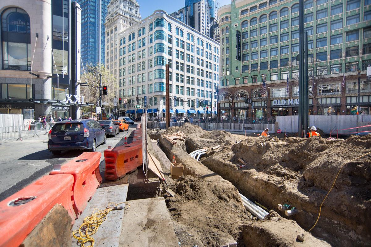 Construction excavation on downtown San Francisco street with buildings and barriers visible.