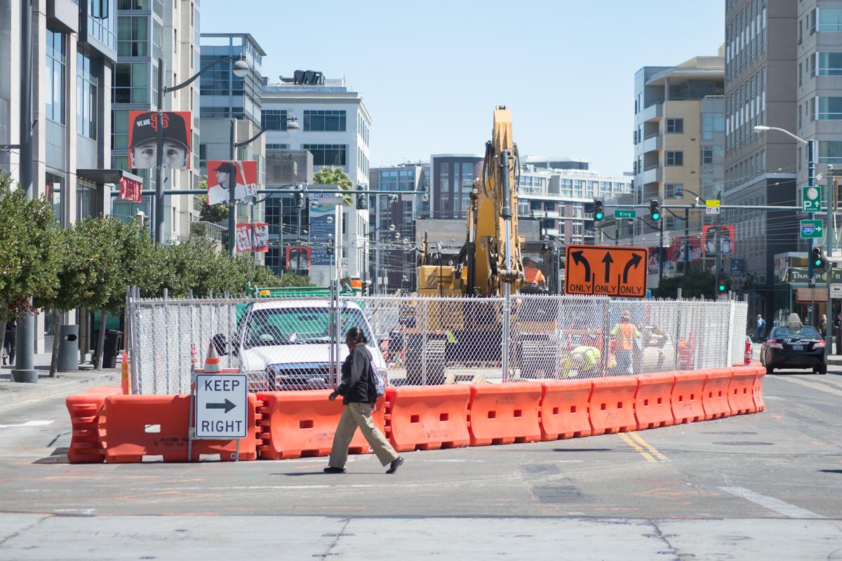 Construction site on 4th Street with orange barriers, excavator, and workers behind chain-link fencing.