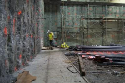 Worker in yellow safety gear at construction site with concrete wall, metal scaffolding, and steel rebar materials.