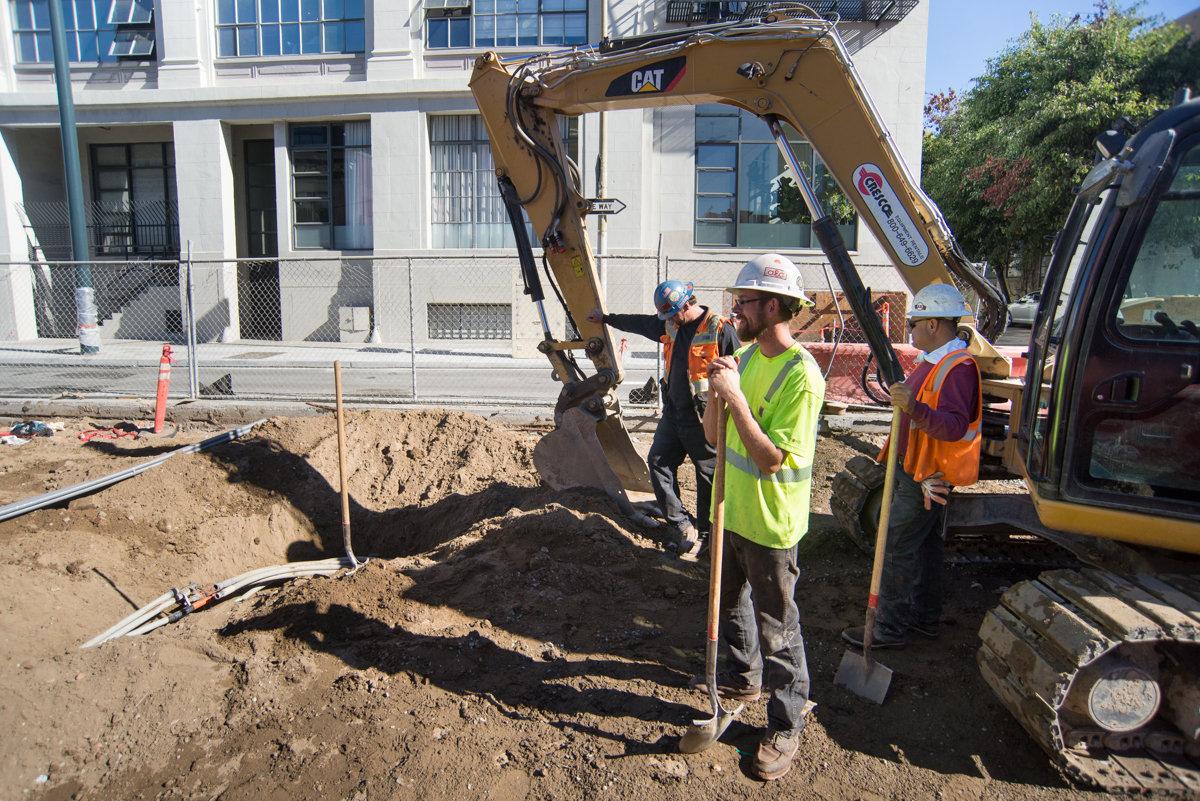 Construction workers and equipment excavating underground utilities on city street near building.