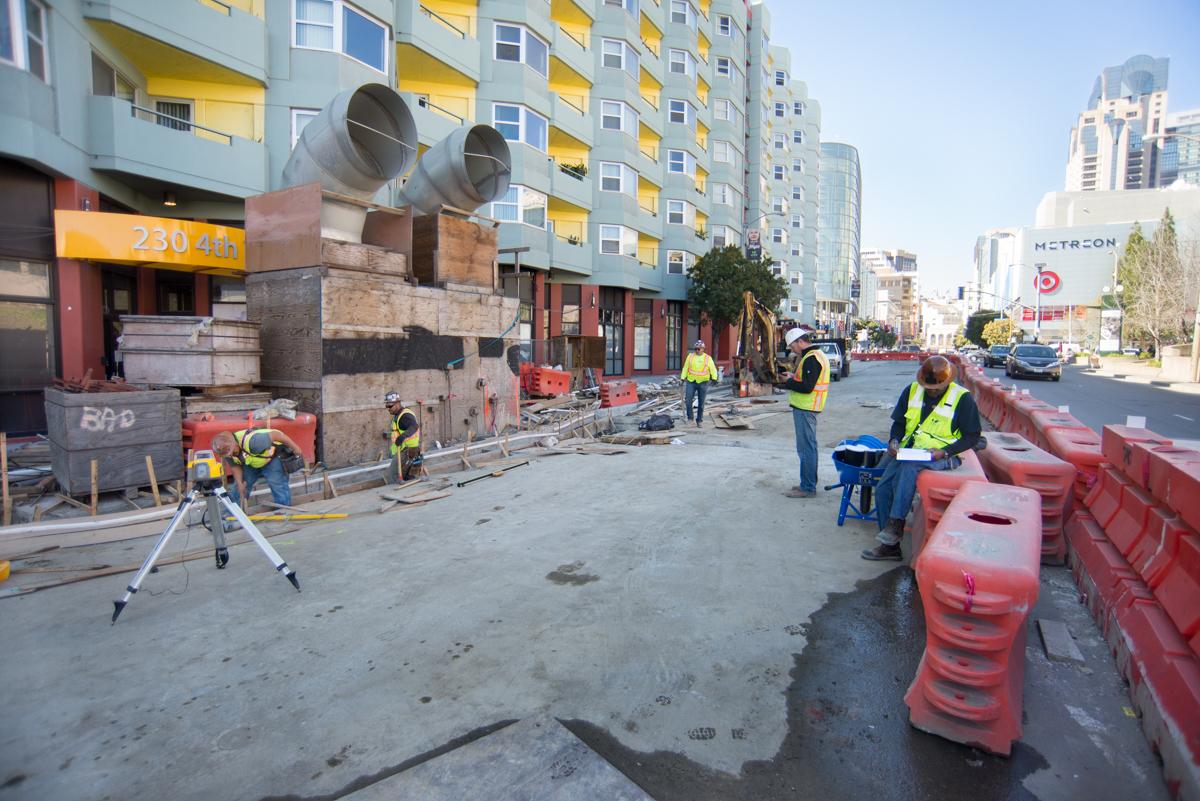Construction workers in safety vests at urban transit station excavation site with red barriers and equipment.