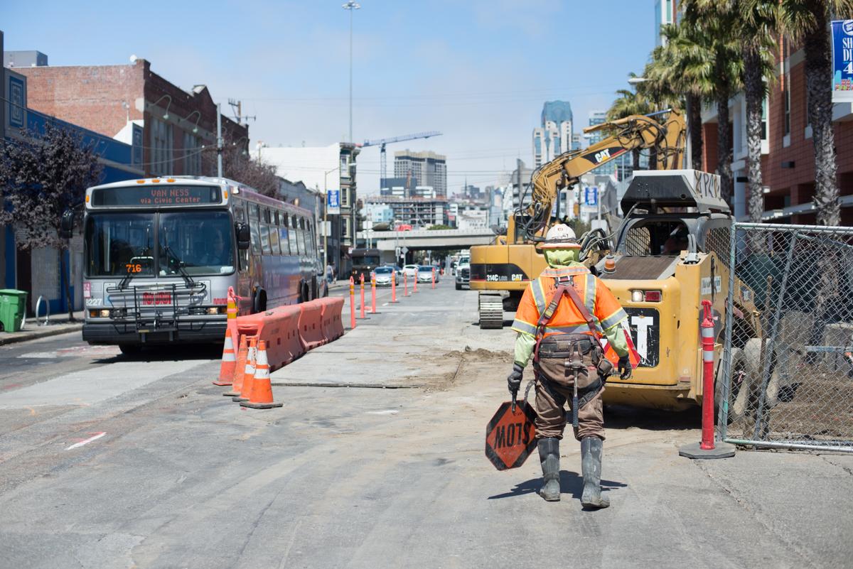 Construction worker directing traffic through street work zone with bus, excavator, and safety barriers visible.
