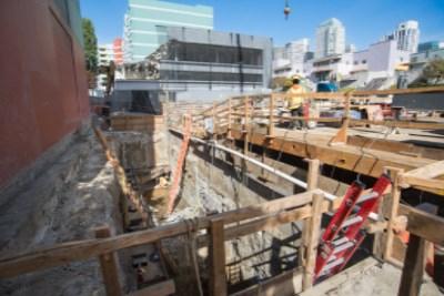 Construction site showing deep trench excavation with wooden support structures and heavy equipment in urban setting.