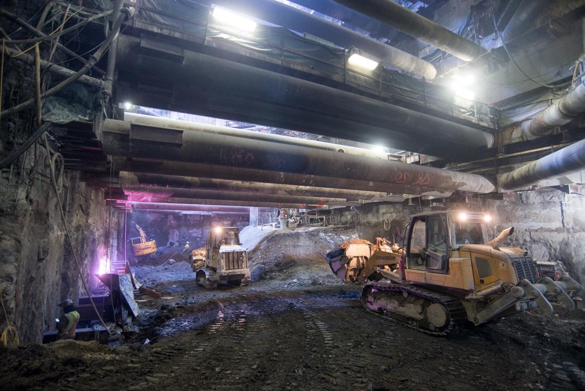 Underground construction site with bulldozers and excavation equipment beneath steel beams and industrial lighting.