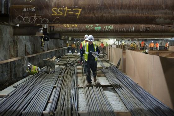 Worker in hard hat and safety vest walking on rebar in underground transit station construction area.