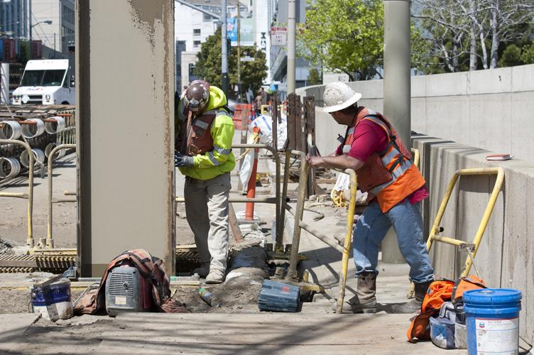 Construction workers in safety vests and hard hats perform headwall construction work on 4th Street using hand tools and equipment.