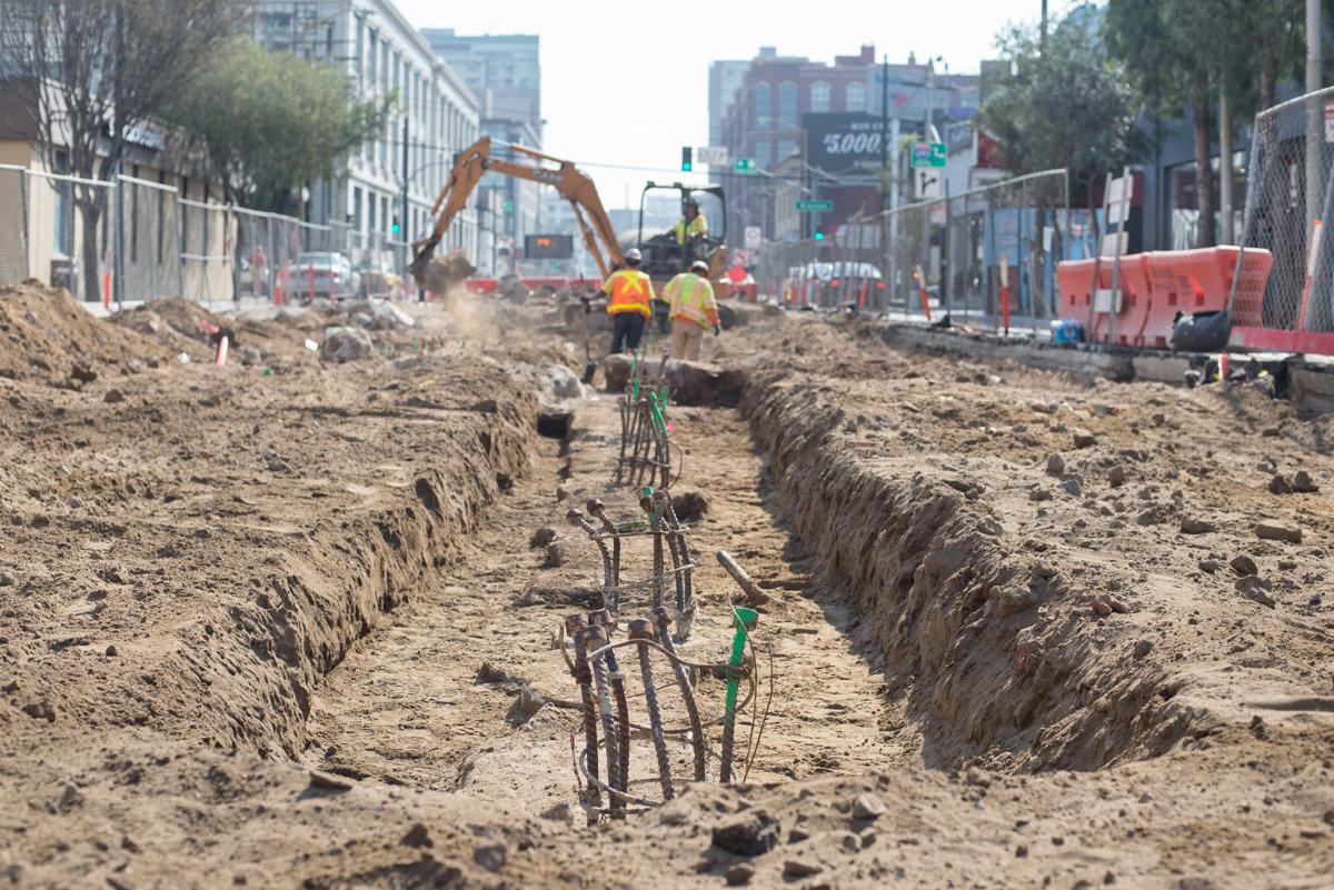 Construction trench with exposed rebar and excavation equipment on urban street.