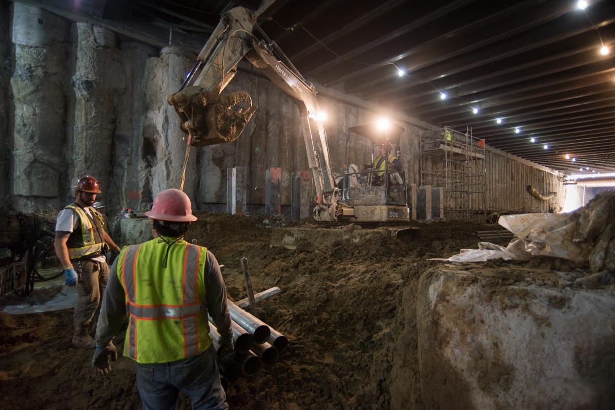 Workers operate excavation equipment in underground transit station construction site beneath street level.
