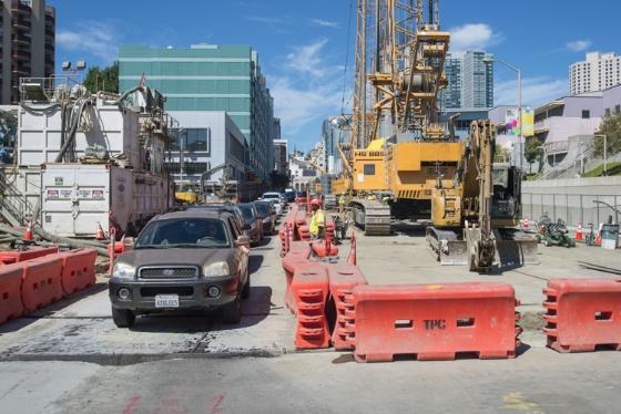 Construction site on 4th Street with one open traffic lane, red barriers, heavy equipment, and workers building roadway walls.