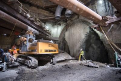 Underground construction site with yellow excavator, concrete pump truck, and large circular tunnel boring equipment in cavern.