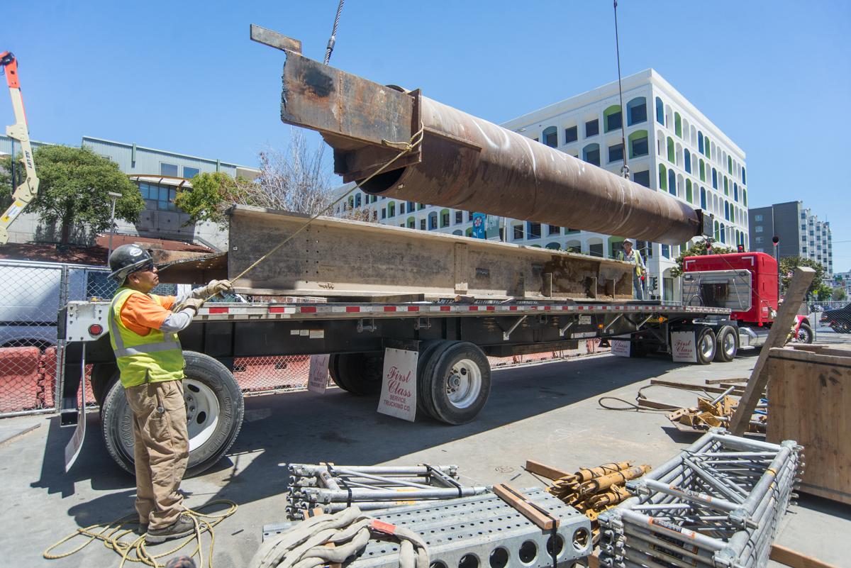 Worker in safety gear guides large steel strut onto flatbed truck at Central Subway construction site.
