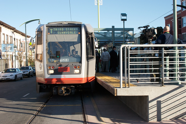 Light rail train stopped at platform with passengers boarding, urban street visible in background.