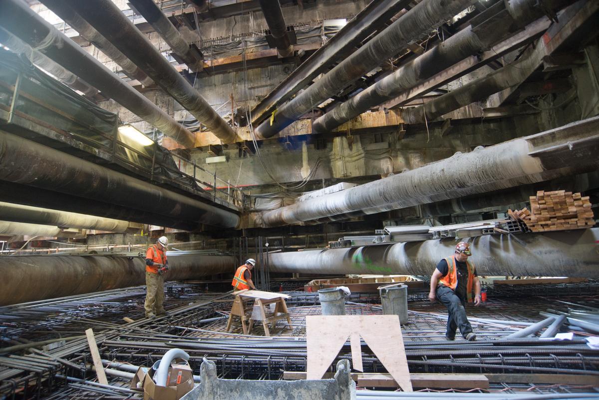Workers in hard hats construct curved concrete forms inside an underground transit tunnel using steel support structures.