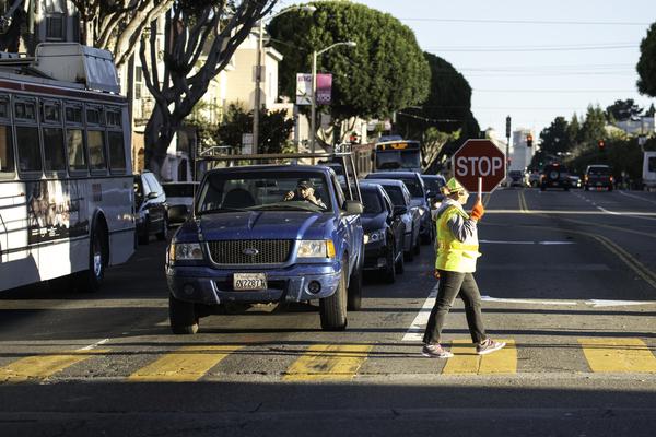 School crossing guard in yellow safety vest directing traffic at pedestrian crosswalk on city street.