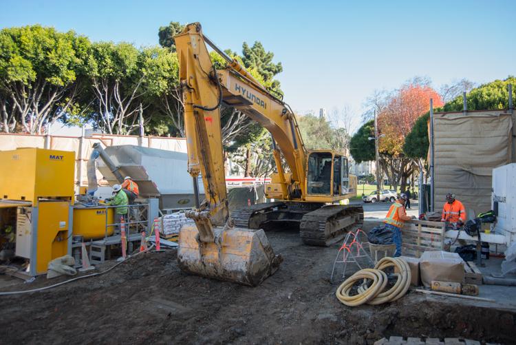 Yellow excavator operates at Powell Street construction site with workers in safety vests and equipment nearby.