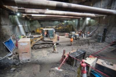 Construction site showing excavated station headhouse with concrete walls, equipment, and workers during underground transit station construction.