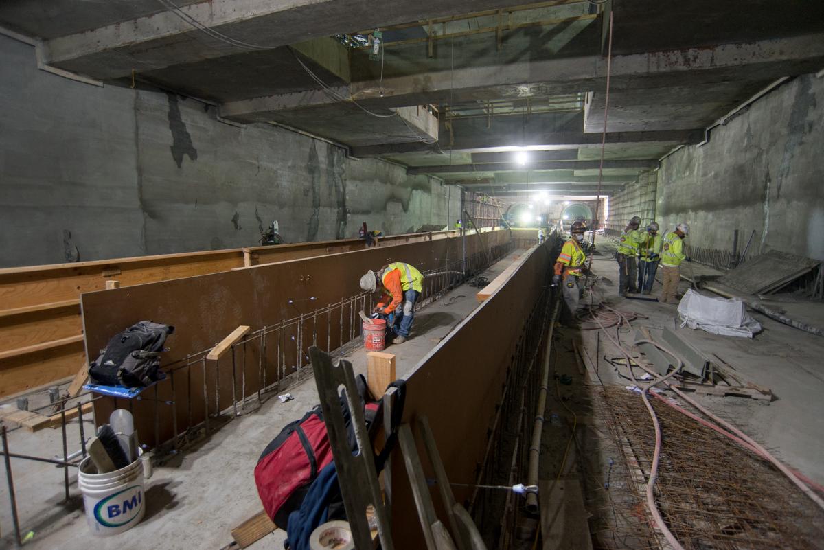 Construction workers in safety vests build concrete forms and wooden platforms inside an underground transit station tunnel.