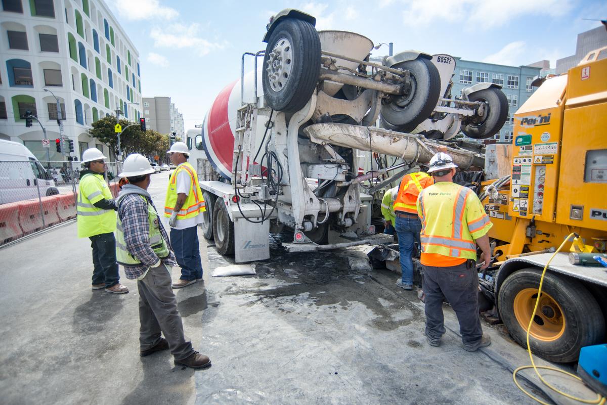 Overturned cement truck with workers and emergency responders at construction site near urban buildings.